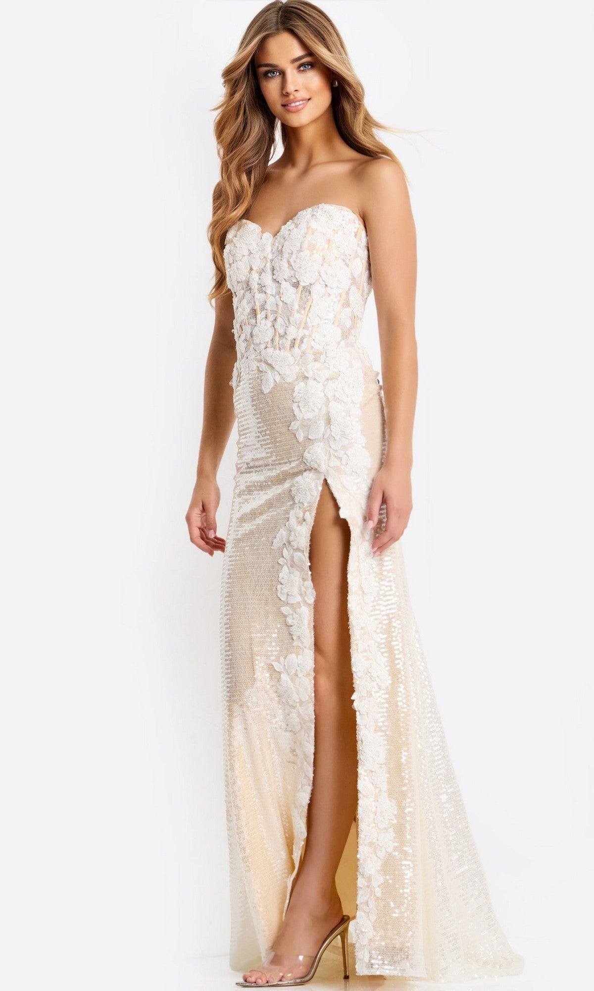 A woman wears the Jovani 49362 long prom dress—a strapless, floor-length cream gown with floral details and a high slit—paired with clear high heels.